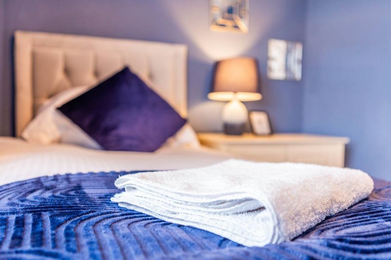 Modern bedroom with neatly arranged towels on bed, purple cushion, bedside lamp and neutral headboard