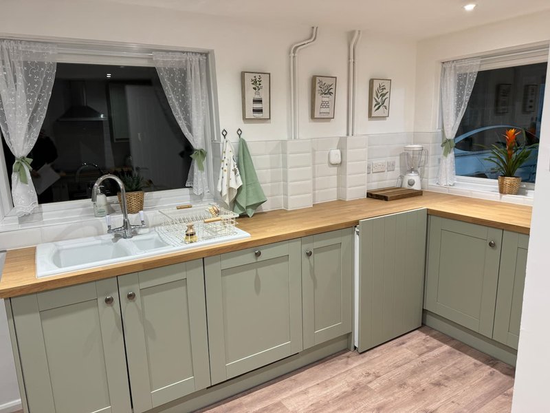 Modern kitchen with sage green cabinetry, wooden countertop, white sink and windows with curtains