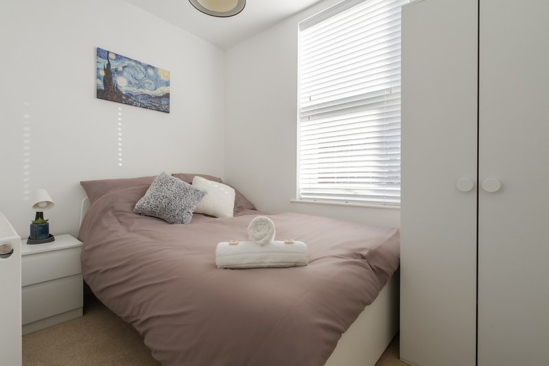 Bedroom with double bed, mauve bedding, white nightstand, framed artwork and window with blinds