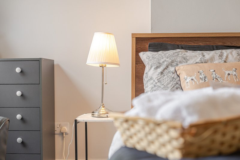 Bedroom with wooden bed frame, bedside lamp, dark drawers and neutral walls