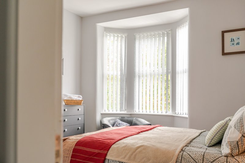 Bright bedroom with double bed, red throw, small chest of drawers and vertical blinds