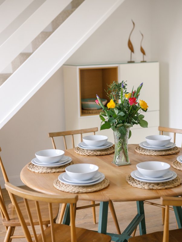 Dining area with round wooden table, place settings, fresh flowers in vase, and modern storage shelving