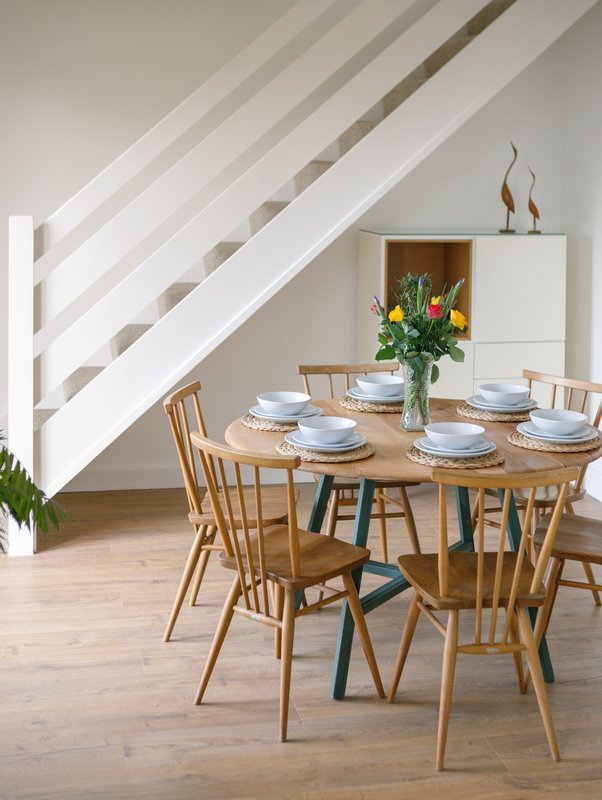 Dining area with wooden table, chairs, place settings, and fresh flowers under modern staircase