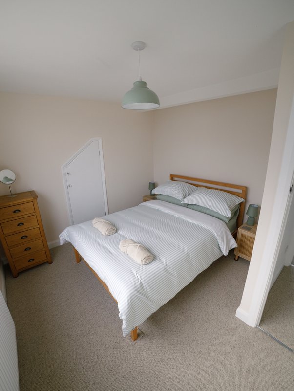 Bedroom with wooden bed frame, white bedding, wooden chest of drawers, and pendant light