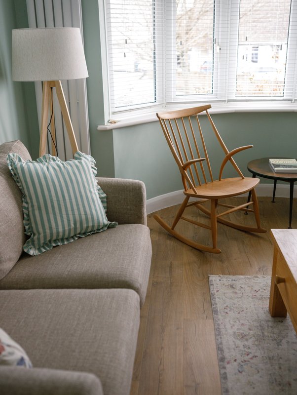 Living room with beige sofa, striped cushion, wooden rocking chair, tripod lamp and windows
