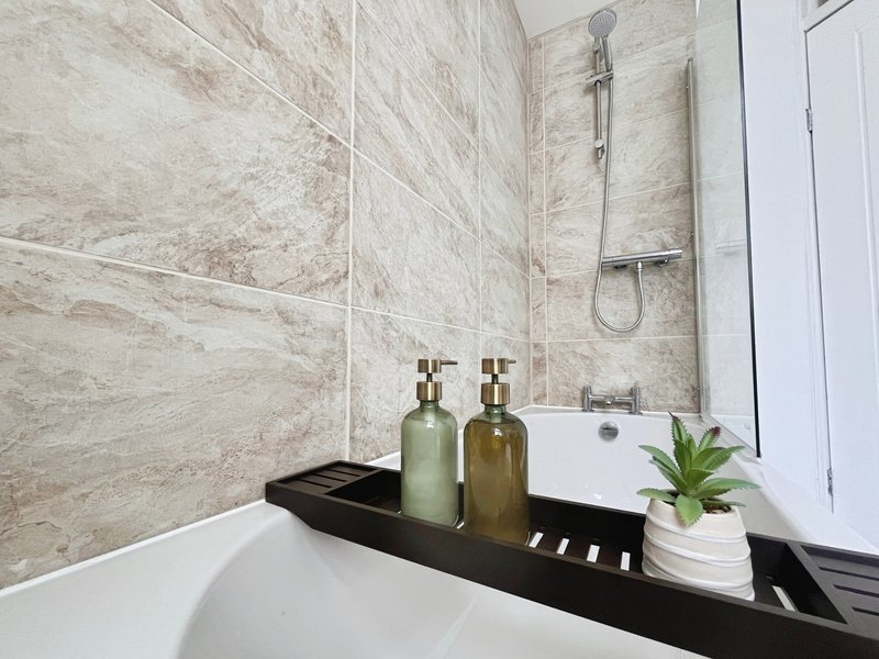 Modern bathroom with marble-effect tiles, white bathtub, and black shelf holding soap dispensers and plant