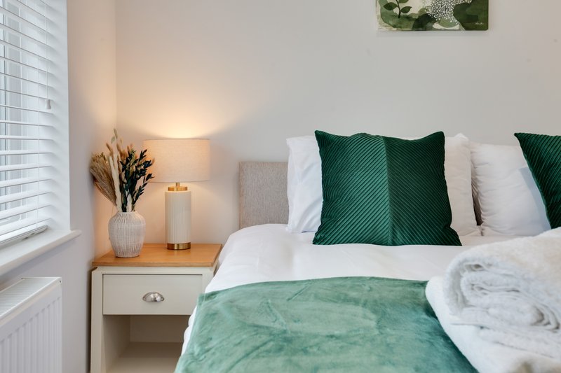 Bright bedroom with double bed, green pillows, white bedding, wooden nightstand and table lamp