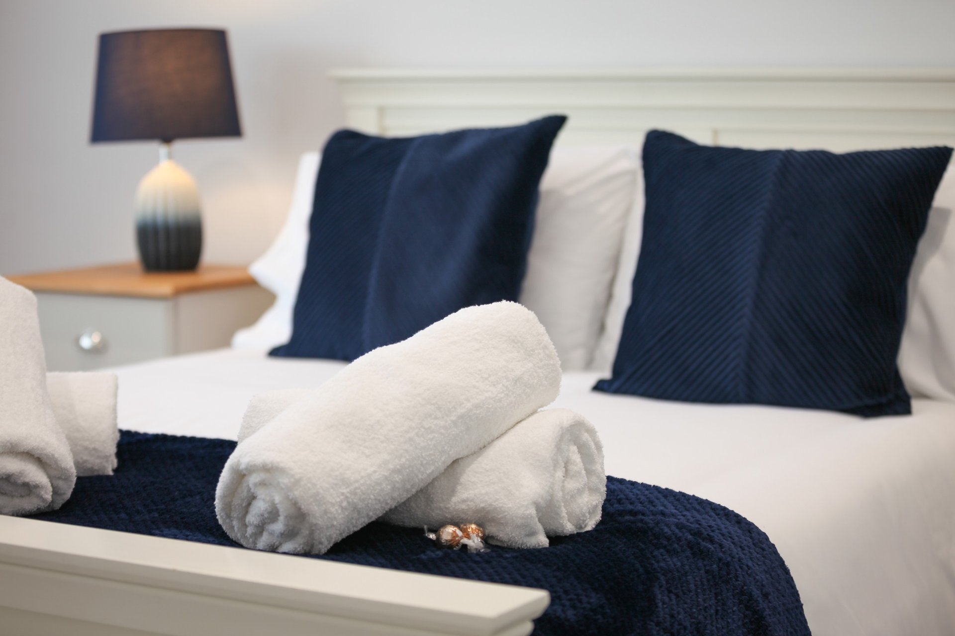Bedroom with white bed, navy and white pillows, rolled towels, and bedside lamp