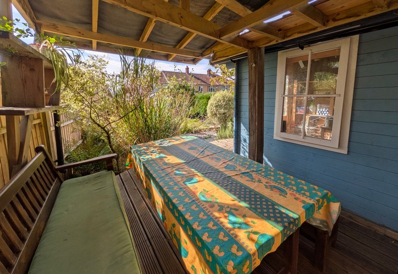 Covered porch with wooden beams, table with colorful cloth, wooden chairs and garden view