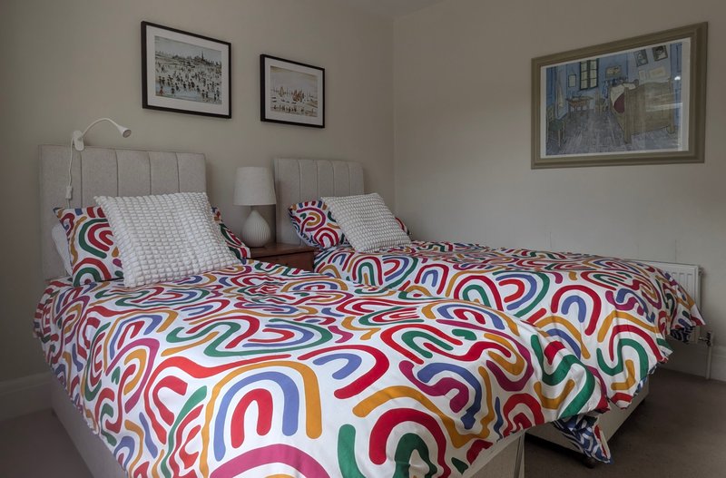Bright bedroom with two single beds, colorful patterned bedding, framed wall art, and bedside lamp