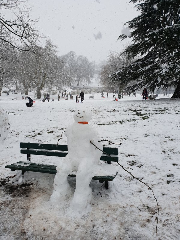 Snowman built in a snowy park with benches, bare trees and distant figures
