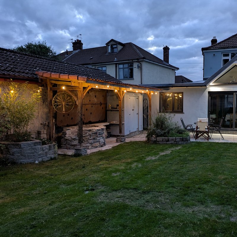 Residential garden at dusk with illuminated wooden pergola, seating area, and house exterior