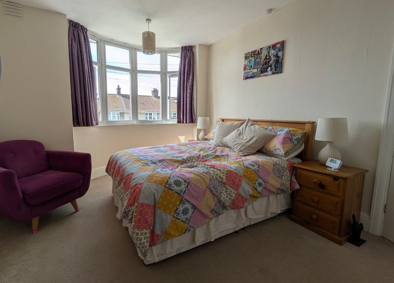 Bright bedroom with double bed, colorful patchwork quilt, purple armchair, wooden nightstand and window with purple curtains
