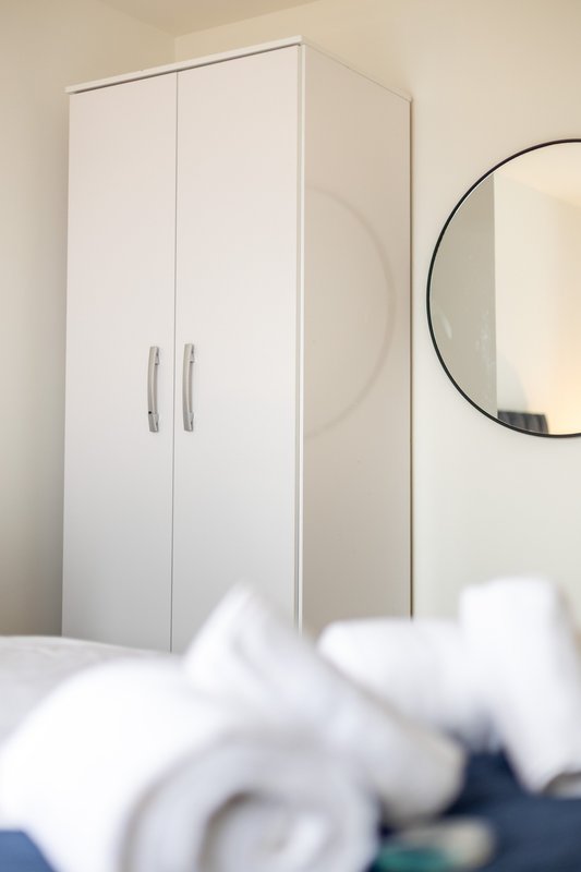 Bedroom corner with white wardrobe, round mirror, and folded towels in foreground