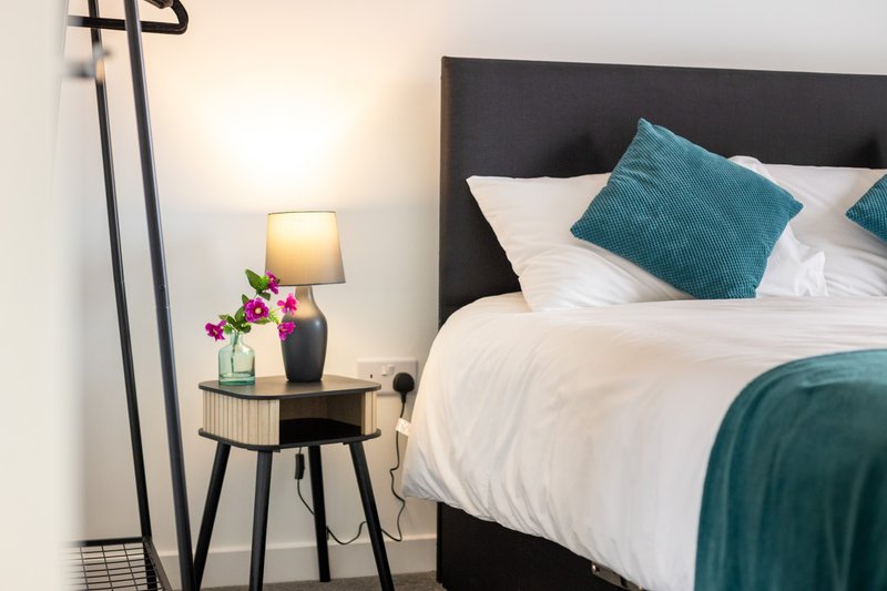 Modern bedroom with black headboard, white linens, teal pillows and throw blanket, bedside table with lamp and flowers