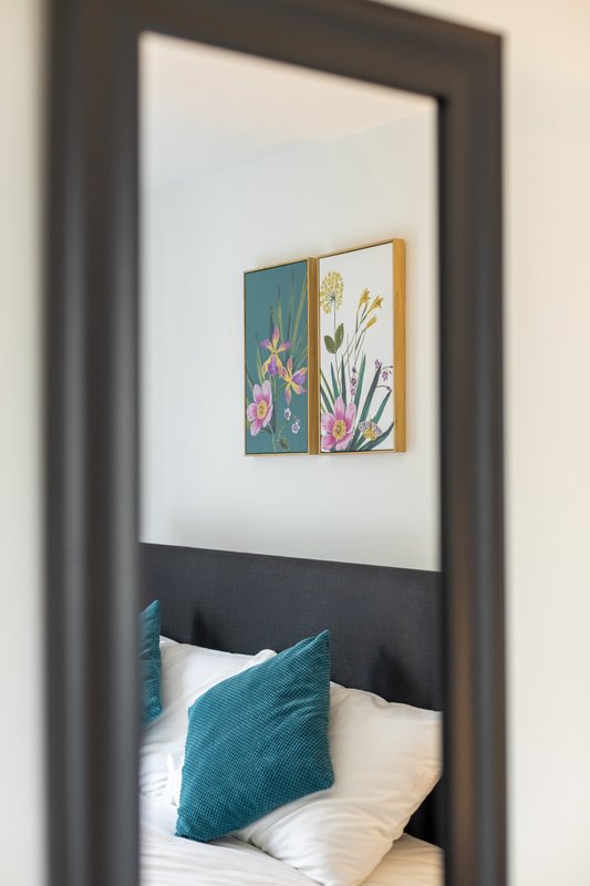 Modern bedroom viewed through doorway with framed floral artwork and teal cushion on bed