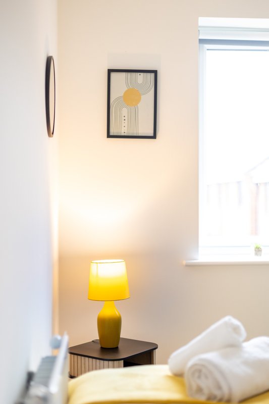 Bedroom corner with yellow table lamp, framed sun artwork, and rolled white towels on bed