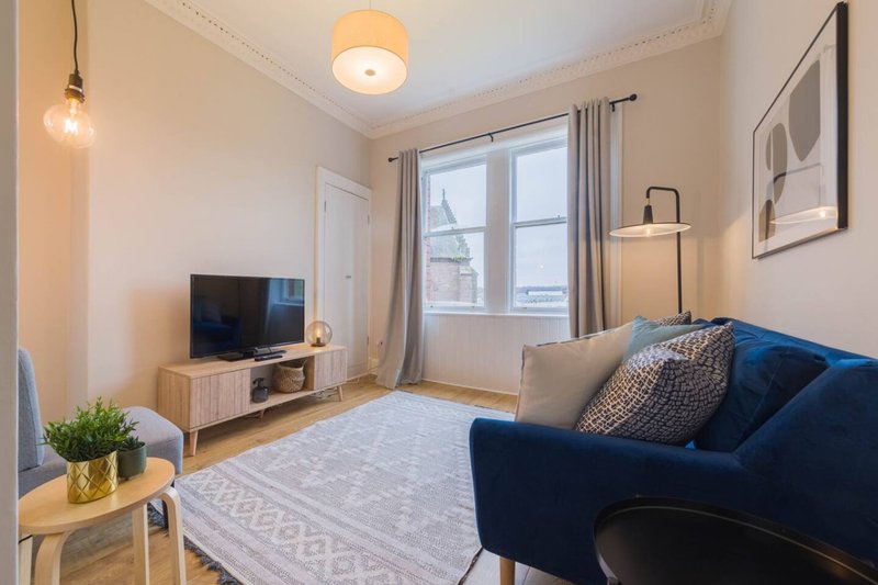 Modern living room with blue sofa, TV stand, pendant lighting, and large windows with city views
