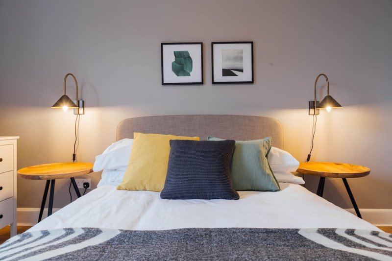 Modern bedroom with gray upholstered headboard, colorful pillows, wooden side tables and brass wall lamps