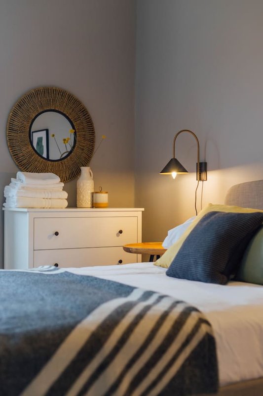 Bedroom with double bed, white bedside table, round woven mirror, wall lamp and folded towels