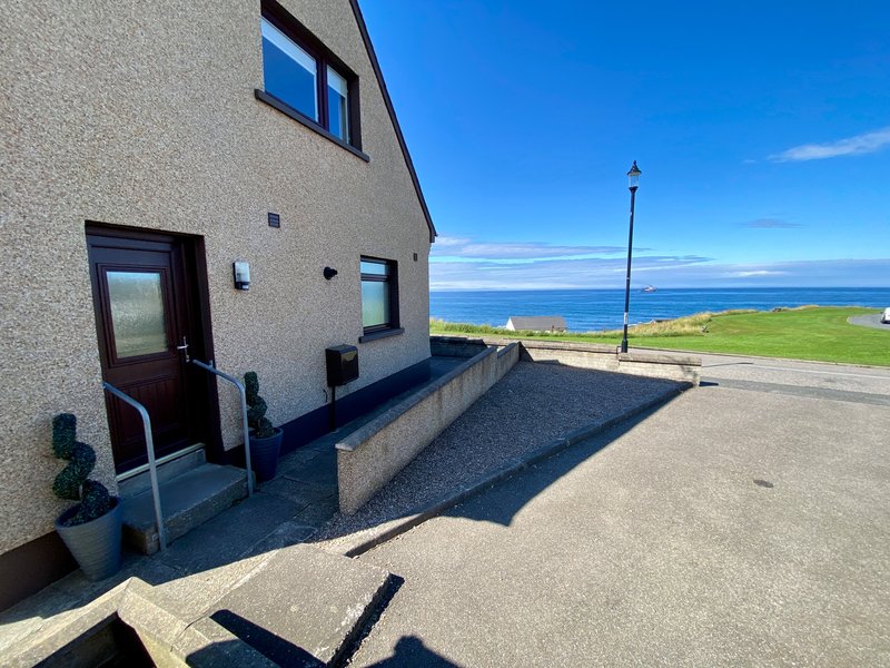 Exterior of stone house with entrance door, driveway, and coastal sea view beyond