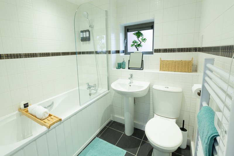 Clean modern bathroom with white tiles, pedestal sink, toilet, bathtub with shower screen and heated towel rail