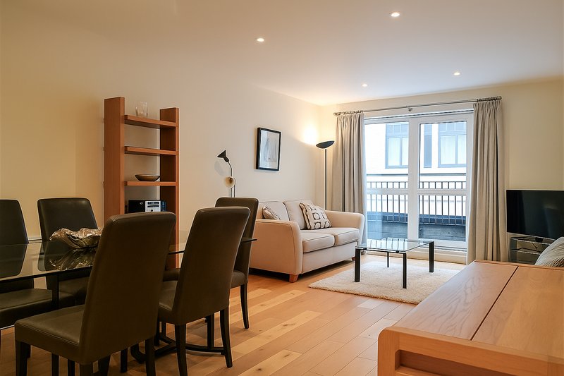 Open-plan living and dining area with wooden flooring, dining table, chairs, sofa, shelving, and glass doors to balcony