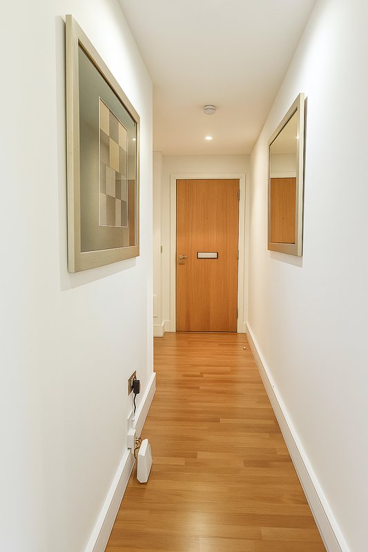 Narrow hallway with wooden floor, white walls, framed wall art, and wooden door