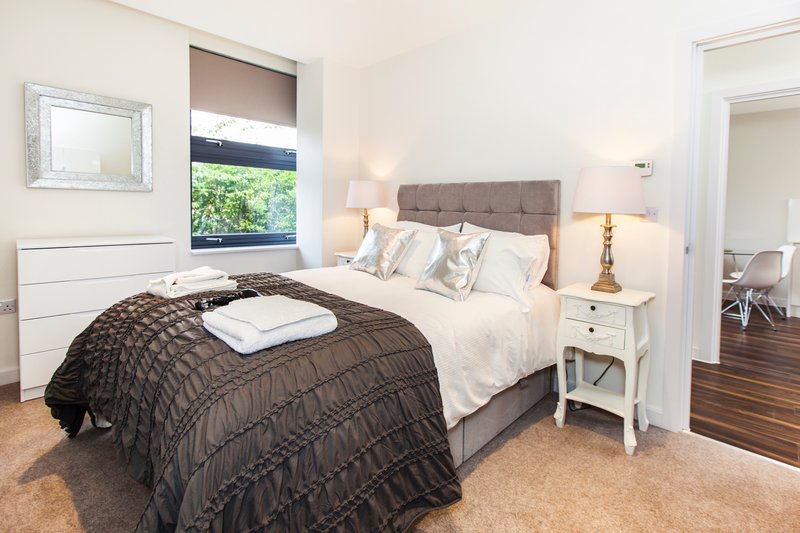 Bedroom with double bed, grey headboard, white bedside table, and window with blinds