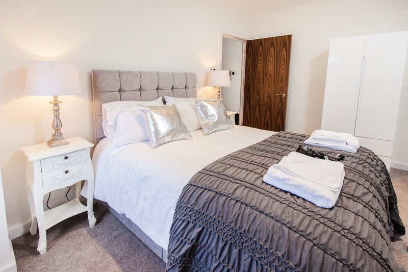 Bright bedroom with double bed, grey upholstered headboard, white bedding and dark throw, wooden wardrobe
