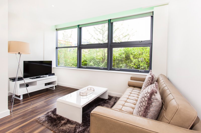 Modern living room with beige sofa, TV stand, floor lamp, and large windows overlooking green trees