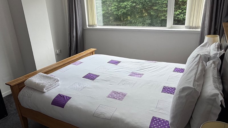 Bedroom with wooden bed frame, white bedding with purple accents, and large window with grey curtains