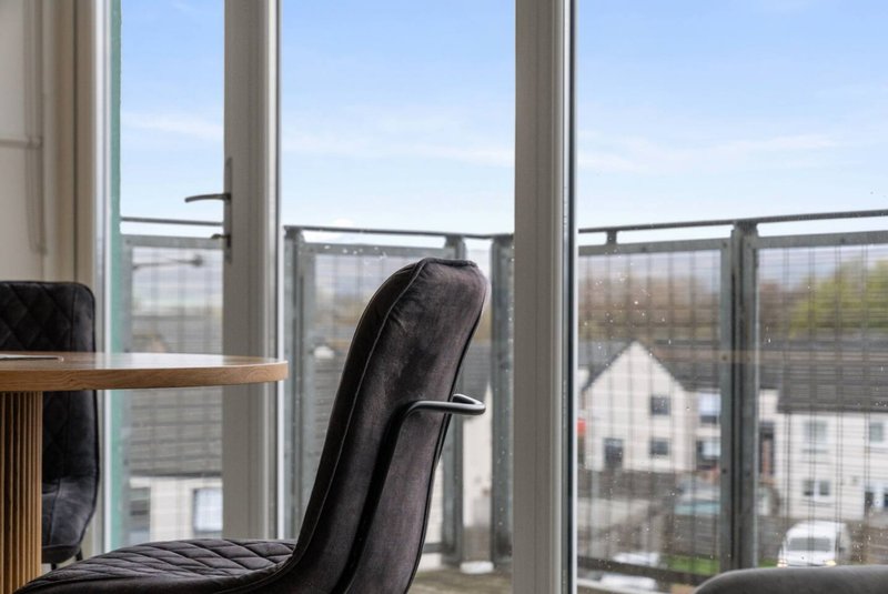 Modern apartment balcony with floor-to-ceiling windows, black office chair, wooden desk, and suburban street view