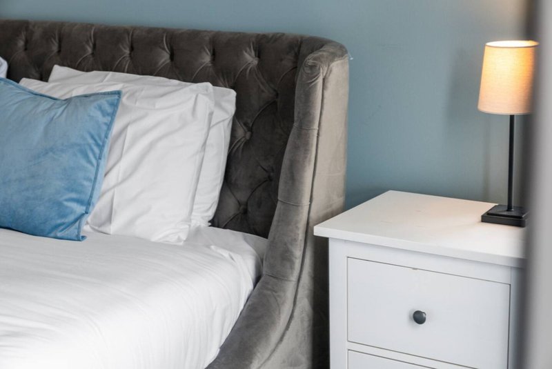 Bedroom with grey upholstered bed, white bedding, blue pillow, white nightstand and lamp.