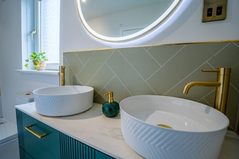 Modern bathroom with dual white vessel sinks, gold fixtures, circular mirror and teal vanity
