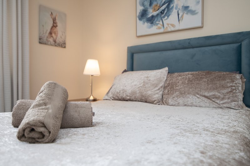 Bedroom with blue upholstered headboard, white bedding, rolled towel, and bedside lamp