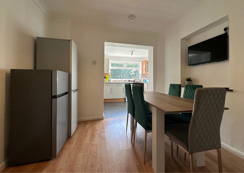 Open-plan kitchen and dining area with refrigerator, wooden table, upholstered chairs and wall-mounted TV