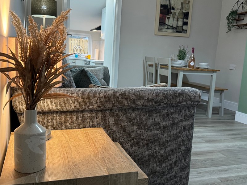 Open-plan living room with gray upholstered sofa, wooden table, dried flower arrangement, and dining area visible in background