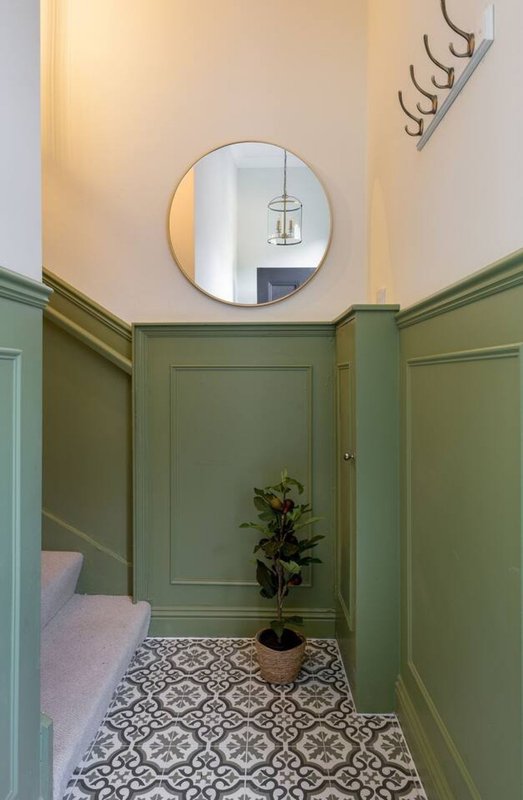 Hallway with sage green panelled walls, round mirror, potted plant and patterned floor tiles