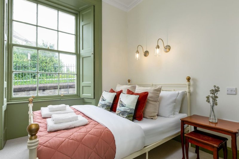 Bright bedroom with double bed, red quilted bedspread, white pillows, green window frame and pendant lights