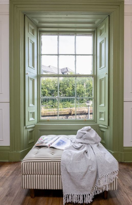 Window seat with green-painted frame, cushioned bench, rumpled bedding and throw blanket, countryside view