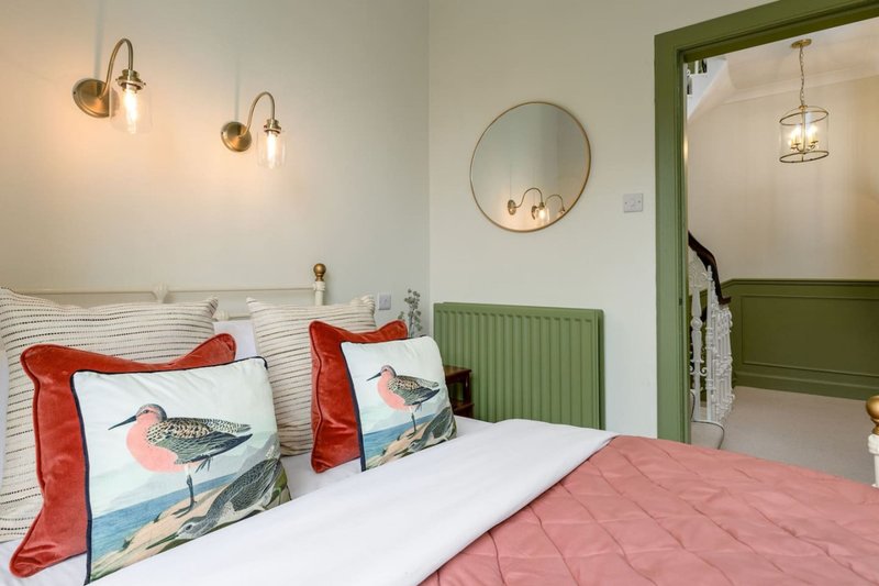 Bright bedroom with double bed, striped headboard, decorative bird pillows, brass wall sconces, and green accent wall