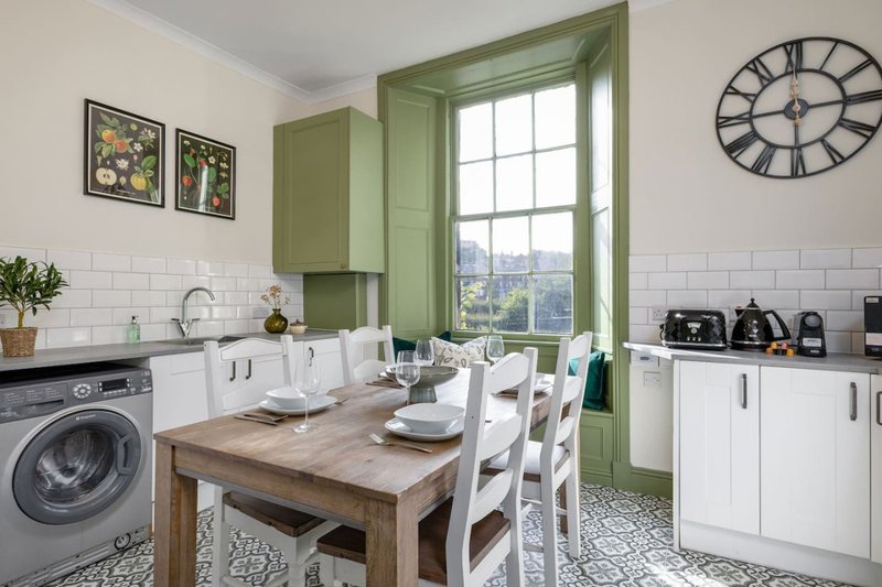 Modern kitchen-diner with white cabinetry, wooden table, washing machine, and green-framed window overlooking cityscape