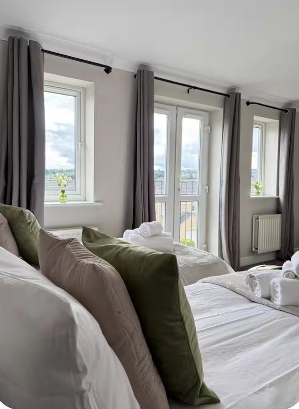 Bright bedroom with double beds, green cushions, grey curtains and balcony doors with countryside views