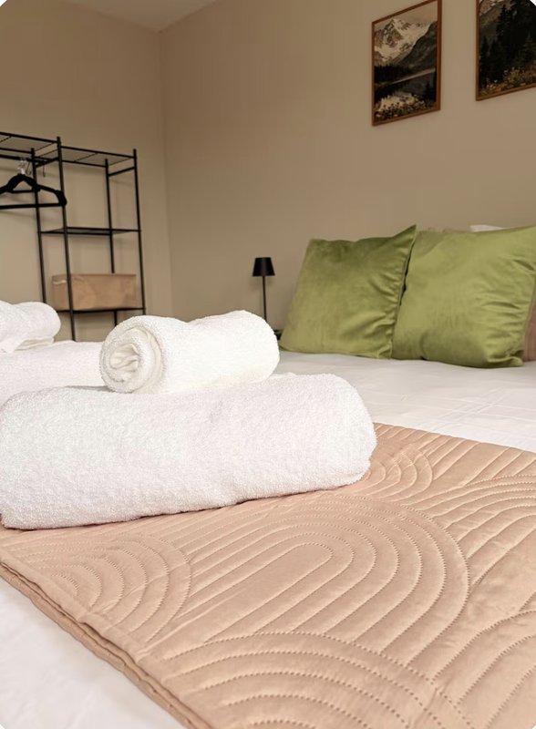 Bedroom with white rolled towels on bed, green cushions, metal bedframe, and framed wall art