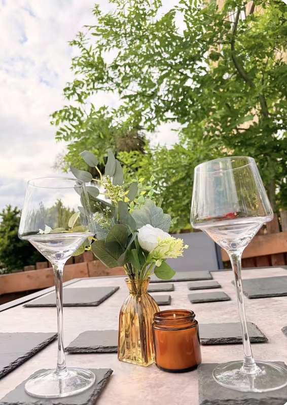 Outdoor dining table with wine glasses, flower vase, and candle under green foliage