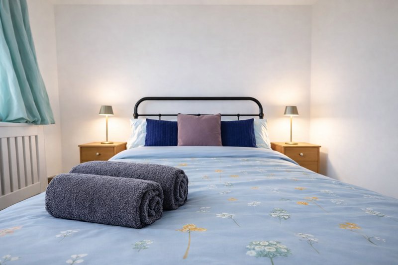 Bright bedroom with double bed, metal frame headboard, blue patterned duvet, decorative pillows and folded towels
