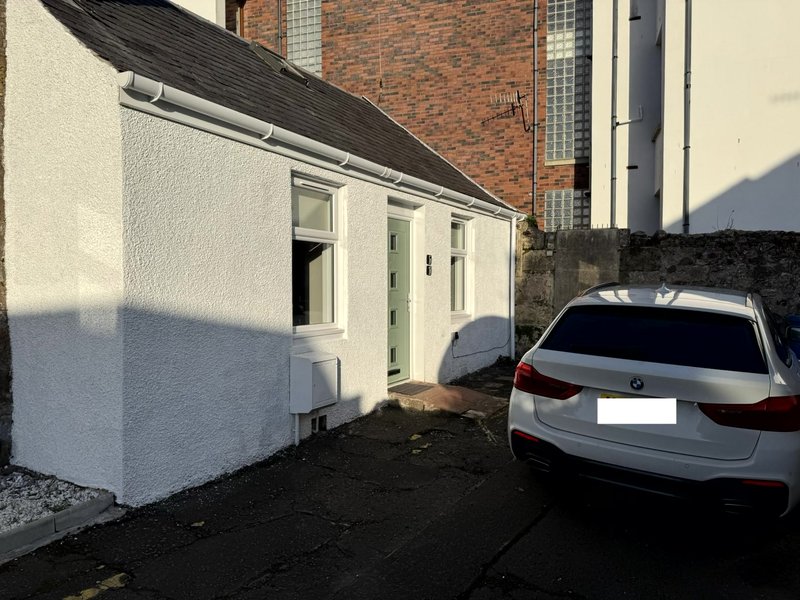 White-rendered single-story cottage with green door and parking, adjacent to brick building