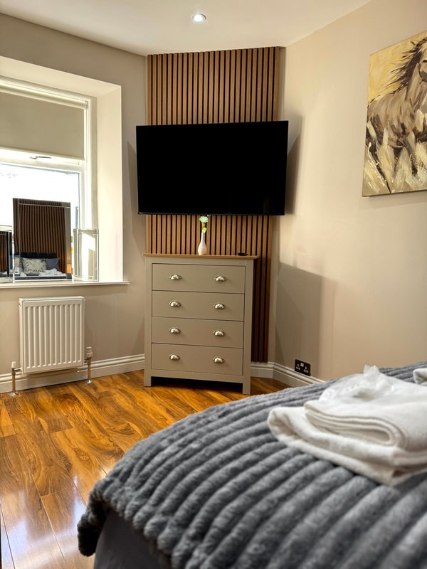 Bedroom with wooden floor, wall-mounted TV, grey dresser, window with curtains, and bed with dark textured bedding