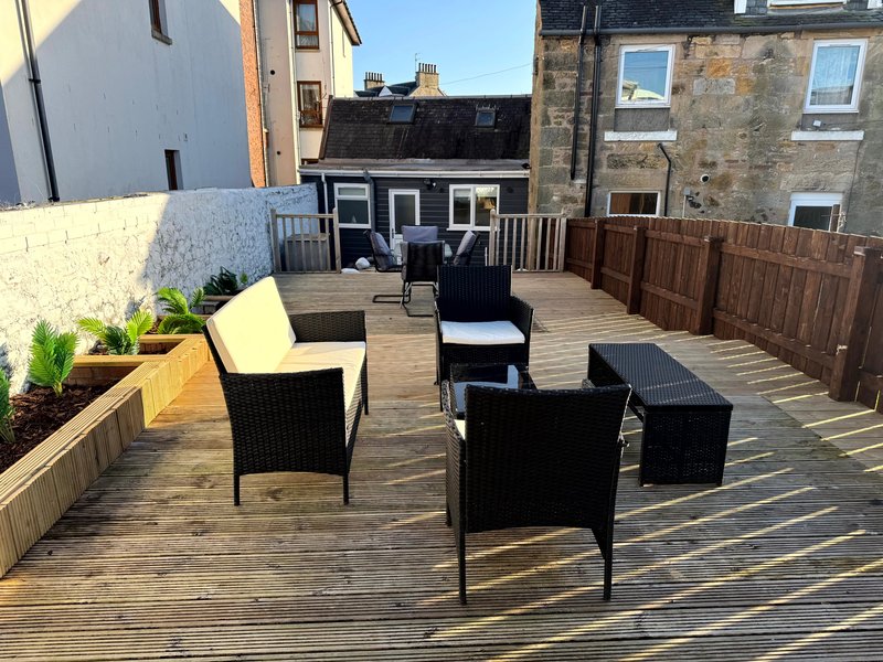 Wooden deck with black rattan garden furniture, planters, and surrounding buildings